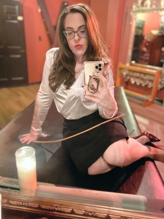Chicago Dominatrix Sophia Chase wearing a white blouse and black pencil skirt seated on her bondage table taking a selfie in an ornate mirror.